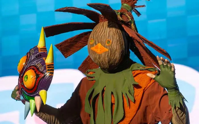 A cosplayer in a torn sailor uniform with a furred monster arm, lifting a fox mask on stage